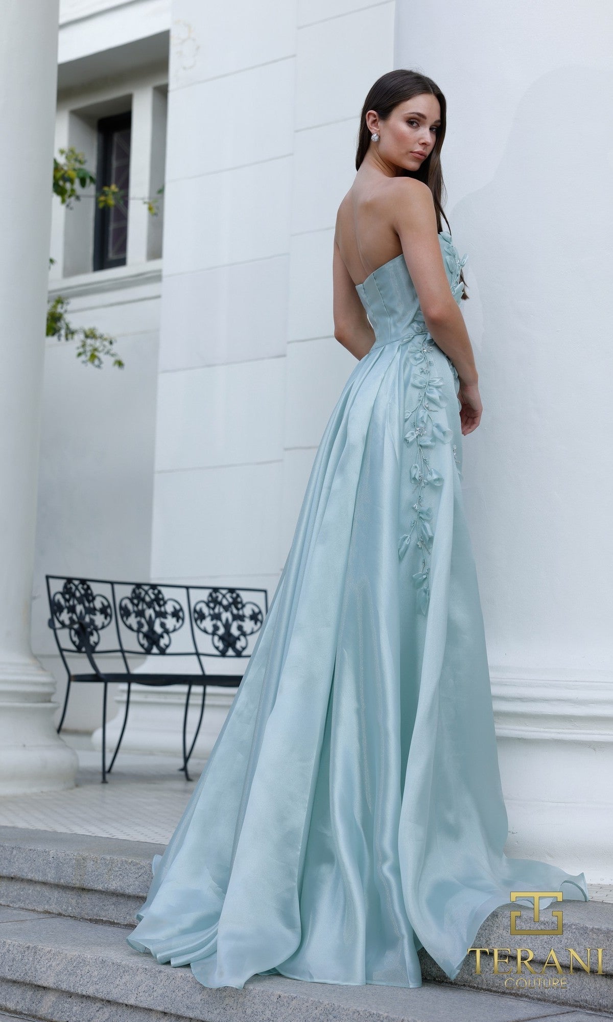 Woman in a light blue, strapless Terani 261E6195 formal dress with floral details stands on stone steps before a white building, looking over her shoulder?a stunning look.